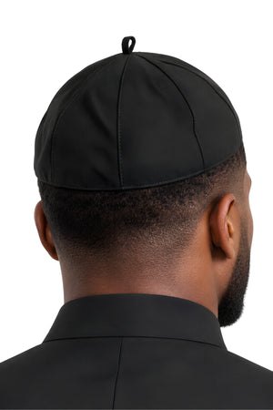 Black clergy zucchetto cap worn on a man's head, showing the fitted skullcap shape, eight-panel construction, and neat lower edge from the back.