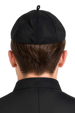 Back view of a person wearing a black clergy zucchetto cap with matching black clerical clothing, showcasing the stitched panel seams, rounded fit, and fabric loop at the crown against a white background.