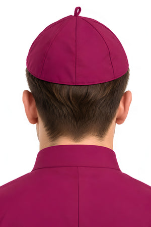 Back view of a person wearing a red clergy zucchetto cap with matching red clerical clothing, showing the fitted cap shape, stitched panel seams, and crown loop.
