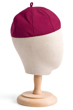Magenta eight-panel clergy zucchetto skullcap displayed on a beige linen head mannequin with a wooden stand, highlighting the stitched seams and crown loop.
